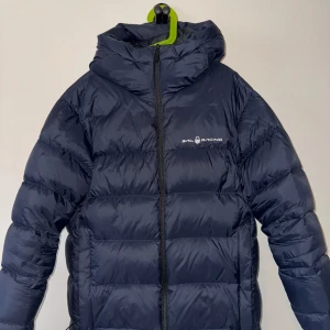 Mörkblå dunjacka från Sail Racing - Säljer en mörkblå dunjacka från Sail Racing med huva och dragkedja. Jackan har en stilren design med Sail Racing-logga på bröstet. Perfekt för kyliga dagar.                 Nypris:2000kr, Säljer för:600kr