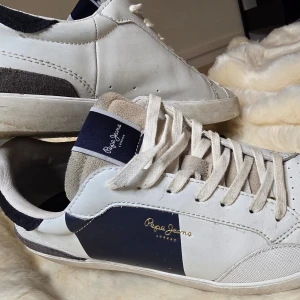 Vita sneakers från Pepe Jeans - Snygga vita sneakers från Pepe Jeans med mörkblå detaljer och snörning. Skorna har en klassisk design med en kontrasterande mörkblå panel på sidan och Pepe Jeans-logga i guld. Perfekta för en stilren look.