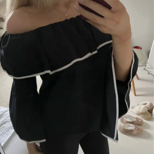 Svart offshoulder blus med vita detaljer - Snygg svart offshoulder blus med vita kanter. Blusen har vida ärmar och en luftig passform, perfekt för en stilren look.