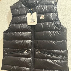 Svart dunväst från Moncler - Snygg svart dunväst från Moncler med dragkedja och två fickor framtill. Västen har en quiltad design och Moncler-logga på bröstet. Perfekt för kyligare dagar. Storlek M