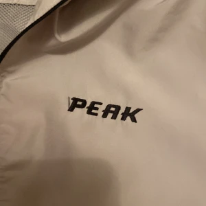 Beige vindjacka från Peak Performance. - Säljer en vit vindjacka från Peak Performance med halv dragkedja och elastiska muddar. Jackan har en broderad logga på bröstet och är perfekt för blåsiga dagar. Den har en klassisk passform och är tillverkad i ett lätt syntetmaterial.