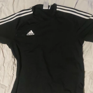 Svart t-shirt från Adidas - Säljer en svart t-shirt från Adidas med de klassiska tre ränderna på axlarna. T-shirten har en rund hals och korta ärmar, perfekt för sport och träning. Adidas-loggan är tryckt på bröstet. Tillverkad i Vietnam.