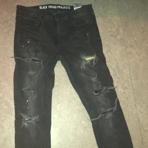 Svarta jeans från Black Squad Projects - Snygga svarta jeans med slitna detaljer och hål för en trendig look. Byxorna har en normal passform och är perfekta för en avslappnad stil. De har en knappgylf och är tillverkade i ett slitstarkt material.