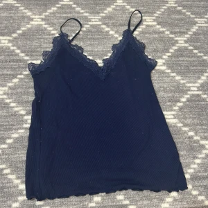 Mörkblå ribbad linnetopp från Amisu - Säljer en stilren mörkblå ribbad linnetopp från Amisu med spetsdetaljer vid halsringningen. Toppen har smala axelband och en v-ringad design som ger en elegant touch. Perfekt för att lägga till en feminin känsla till din outfit.