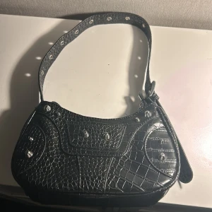 Svart axelväska med krokodilmönster - Snygg svart axelväska med krokodilmönster och silverdetaljer. Väskan har en dragkedja på baksidan och en justerbar axelrem med nitar. Perfekt för att ge din outfit en edgy touch. Finns också två fickor på insidan och en på baksidan🥰