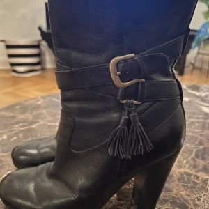 Svarta skinnstövlar med tofsdetalj - Snygga svarta stövlar i skinn med en dekorativ tofs och spänne på sidan. De har en klack som ger extra höjd och stil. Perfekta för att komplettera en trendig look.