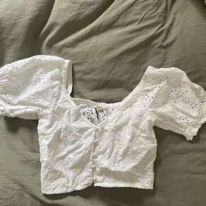 Vit blus från Nelly - Säljer en söt vit blus från NLY Trend med broderade blommor och knappar framtill. Blusen har korta puffärmar och en v-ringad design. Perfekt för en somrig look! 🌸