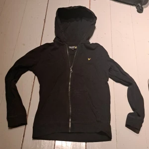 Svart hoodie från Lyle & Scott - Säljer en stilren svart hoodie från Lyle & Scott med dragkedja och huva. Tröjan har långa ärmar och en liten gul logotyp på bröstet. Perfekt för en avslappnad look. Vi har djur hemma. Köparen står för frakt