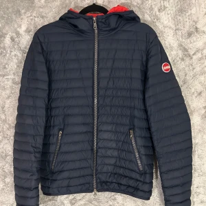 Mörkblå pufferjacka från Colmar - Snygg mörkblå pufferjacka från Colmar med röd insida och huva. Jackan har dragkedja framtill och två fickor med dragkedjor. Perfekt för kyligare dagar med sin quiltade design. Storlek 168