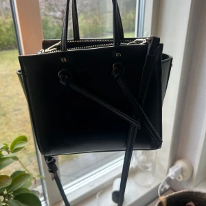 Svart axelväska med dragkedja - Snygg svart axelväska i skinn med praktisk dragkedja upptill. Väskan har en stilren design med två handtag och en justerbar axelrem. Perfekt för att bära dina dagliga nödvändigheter med stil.