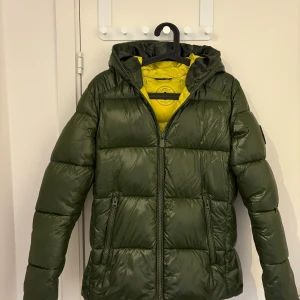 Grön pufferjacka från Save The Duck - Säljer en grön pufferjacka från Save The Duck i storlek S. Jackan har en quiltad design med dragkedja och huva. Insidan är gul vilket ger en snygg kontrast. Perfekt för kyliga dagar!