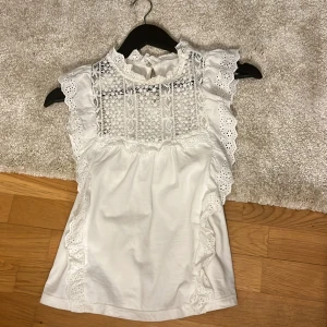 Vit spetsblus - Säljer en vacker vit blus med spetsdetaljer och volanger. Blusen är ärmlös och har en hög hals med knytning i nacken. Perfekt för en elegant look.