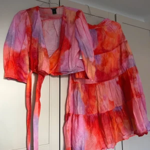 Färgglad omlott topp och kjol  - Säljer en färgglad set från Lindex,  toppen och kjolen har ett gulligt mönster i rosa, lila och orange toner. Blusen är puffärmad med knytband i midjan. Perfekt för sommaren!