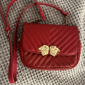 Röd quiltad axelväska med lejonhuvud - Snygg röd axelväska i quiltat mönster med guldfärgade lejonhuvuden som detalj. Väskan har en justerbar axelrem och en praktisk storlek för vardagsbruk. Perfekt för att ge en touch av elegans till din outfit.