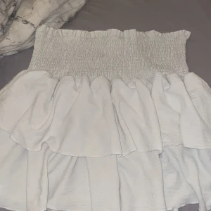 Vit volangkjol - Säljer en söt vit kjol med volanger och smockdetaljer upptill. Perfekt för varma dagar och ger en romantisk touch till din outfit. Kjolen har en kort längd och är lätt att matcha med olika toppar.