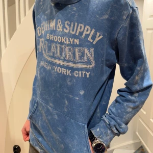 Blå hoodie från R. Lauren - Säljer en blå hoodie från R. Lauren med texten 'Denim & Supply Brooklyn New York City' på framsidan. Tröjan har en unik tvättad look och en stor ficka framtill. Perfekt för en avslappnad stil.