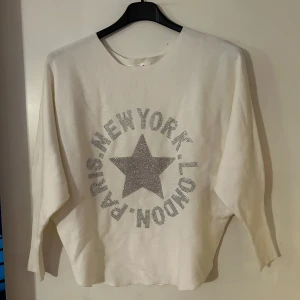 Vit tröja med stjärndetalj onesize - Säljer en vit tröja med en glittrig stjärna och texten 'Paris, New York, London' i silver på framsidan. Tröjan har en avslappnad passform och trekvartsärmar. Perfekt för en trendig look! Helt ny. Storlek onesize.