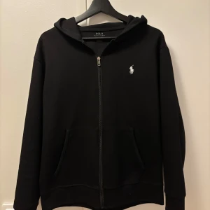 Svart hoodie från Polo Ralph Lauren - Säljer en stilren svart hoodie från Polo Ralph Lauren med dragkedja och det klassiska logotypbroderiet på bröstet. Perfekt för en avslappnad look. Hoodien har långa ärmar och en praktisk ficka framtill. Dock saknar den tyvär dragsko.