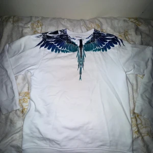Vit tröja med vingmönster från Marcelo Burlon - Säljer en vit tröja från Marcelo Burlon med ett coolt vingmönster i blått och svart runt halsen. Tröjan har långa ärmar och är perfekt för en stilren look. Passar bra till både jeans och sneakers.
