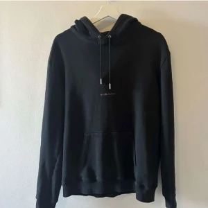 Svart hoodie från Saint Laurent - Snygg svart hoodie från Saint Laurent med diskret logga på bröstet. Tröjan har en klassisk design med dragsko i huvan och en stor ficka framtill. Perfekt för en stilren look. Tyvärr så har snöret tappats bort men ändå en stilren Look, passar S/M men mer åt M hållet 