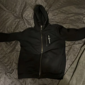 Svart sailracing  hoodie  med dragkedja - Säljer en svart hoodie med dragkedja och huva. Den har en enkel design med ett litet tryck på bröstet. Perfekt för en avslappnad stil.