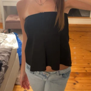 Svart axelbandslös topp - Säljer min snygga svarta festtopp. Den har en fin design med en lätt utsvängd passform. Toppen är enkel att matcha med både jeans och kjol för en chic look. Obs den har ett litet hål, kan skicka bild privat☺️