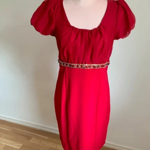 Röd klänning med pärldetaljer - Snygg röd klänning med korta puffärmar och en dekorativ pärlrad i midjan. Perfekt för en festlig kväll. Klänningen har en smickrande passform och är tillverkad i ett lätt och luftigt material.