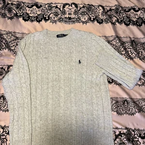 Grå stickad tröja från Ralph Lauren - Säljer en stilren grå stickad tröja från Ralph Lauren med kabelstickat mönster. Tröjan har långa ärmar och en klassisk rund halsringning. Perfekt för kyligare dagar.