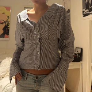 Randig skjortblus  - Snygg randig sljortblus med unik design. Blusen har långa ärmar och en v-ringad krage. Perfekt för en stilren outfit.