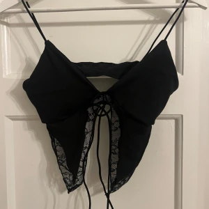 Svart spetsdetaljerad topp - Snygg svart topp med tunna axelband och spetsdetaljer. Toppen har en knytning framtill och en öppen design som ger en elegant look. Perfekt för en kväll ute! Aldrig använd❤️