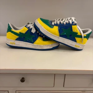 Bapestas ”Brazil” - Snygga och unika sneakers från Bape i en färgglad kombination av blått, gult och grönt. Skorna har en stjärndetalj på sidan och vita snören. Perfekta för dig som vill sticka ut med din stil. Dessa är i storlek 42 och har använts kanske 5-6 gånger. Allt ingår vid köp stickers, extra laces, dustbag samt låda. Dessa är äkta, köpa på bape butiken i Paris. Skicka dm vid intresse!