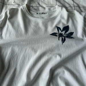 Vit t-shirt med blomtryck från Acne Studios - Säljer en stilren vit t-shirt från Acne Studios med ett snyggt blommönster på bröstet. Perfekt för en minimalistisk look. Tröjan är använd, trycket på baksidan har blivit defekt. Storlek S men sitter som M.