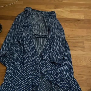 Blå mönstrad klänning - Säljer en blå klänning med ett vitt mönster. Klänningen har långa ärmar och en volangdetalj framtill. Perfekt för en stilren look.