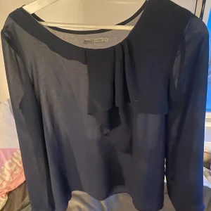 Mörkblå blus med volang - Elegant mörkblå blus från Backed med en stilren volangdetalj framtill. Blusen har långa ärmar och en rund halsringning, perfekt för en chic look.