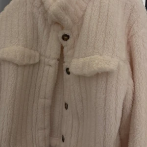 Beige teddyjacka med knappar - Mysig beige teddyjacka med knappar framtill. Jackan har en fluffig textur och är perfekt för kyligare dagar. Den har en avslappnad passform och långa ärmar. Storlek 36 men stor i storleken så passar 38! 