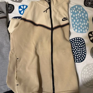 Beige och grå jacka från Nike - Säljer en snygg jacka från Nike i beige och grått. Jackan har en dragkedja framtill och ett stilrent mönster med svarta och bruna ränder över bröstet. Perfekt för en sportig look!