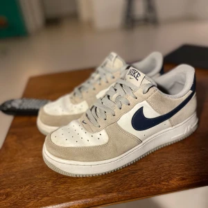 Nike Air Force i beige och vit - Snygga Nike Air Force sneakers i beige och vit med mörkblå detaljer. Skorna har en klassisk design med snörning och perforerade detaljer på tån. Perfekta för en stilren look. Och otroligt sköna. Ej använda så mycket. Tveka inte på att höra av er vid frågor. 