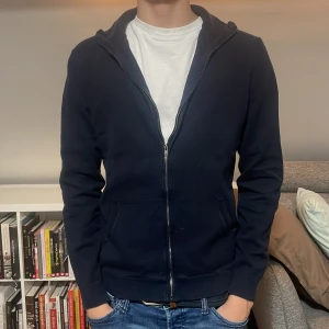 Helzip mörkblå tröja - Säljer en stilren mörkblå hoodie med dragkedja framtill. Den har en klassisk design med långa ärmar och ribbade muddar vid ärmslut och nederkant. Perfekt för en avslappnad look. Tröjan är i 100% bomull stickat material och passar er som är 165-172 ish och hör av er vid frågor och funderingar!