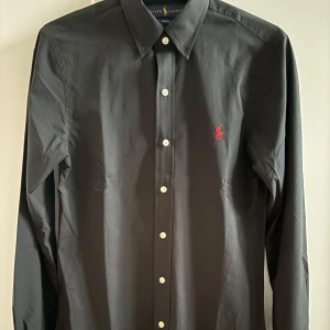 Svart skjorta från Ralph Lauren - Snygg svart skjorta från Ralph Lauren i slim fit. Skjortan har långa ärmar och knappar framtill. Den är dekorerad med en liten röd broderad logotyp på bröstet. Perfekt för en stilren look.