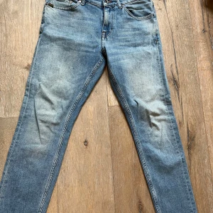 Blå jeans från Tiger of Sweden - Snygga blå jeans från Tiger of Sweden med en klassisk femficksdesign. De har en dragkedja och knapp framtill samt en läderpatch med logotyp baktill. Perfekta för en stilren look. Storlek 29/30
