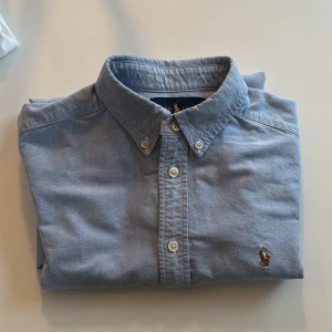 Blå skjorta från Ralph Lauren - Snygg blå skjorta från Ralph Lauren med klassisk krage och knappar framtill. Den har en broderad logga på bröstet som ger en stilren touch. Perfekt för en avslappnad men ändå uppklädd look.