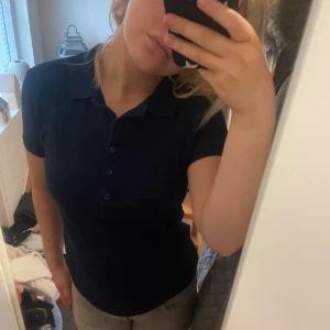 Mörkblå polotopp - Säljer en stilren mörkblå polotopp med korta ärmar och knappar framtill. Perfekt för en avslappnad look. Passar bra till både jeans och kjol.