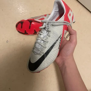 Nike mercurial vapor 15 academy från stadium - Säljer ett par vita och röda fotbollsskor från Nike med svarta detaljer. Skorna har snörning och en modern design med en slitstark sula för optimalt grepp på planen. Perfekta för fotbollsspelare som vill ha stil och funktionalitet.