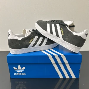 Adidas Gazelle  - Snygga grå Adidas Gazelle sneakers med vita detaljer och klassiska tre ränder. Skorna har en stilren design med snörning och är tillverkade i mocka. Storlek 40 2/3 eller UK 7. Helt nya, aldrig använda. Skriv vid funderingar ✅