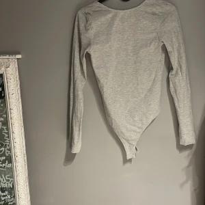 Grå långärmad bodysuit - Snygg grå långärmad bodysuit med djup rundad urringning. Perfekt för lager-på-lager eller att bära som den är. Enkel och stilren design som passar till många olika outfits.