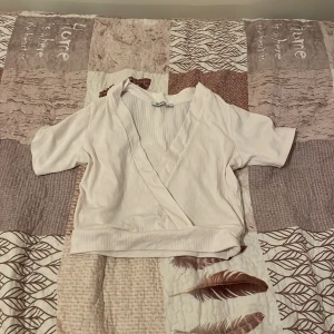 Vit omlottopp - Säljer en stilren vit omlottopp med korta ärmar. Toppen har en v-ringad design och ribbade detaljer vid ärmslut och nederkant. Perfekt för en avslappnad och chic look.