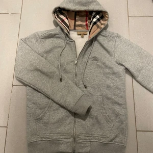 Burrbery hoodie - Säljer en stilren grå hoodie från Burberry med dragkedja och klassiskt rutigt mönster i huvan.Tröjan har långa ärmar och en broderad logga på bröstet. Perfekt för en avslappnad och trendig look. Köpte den på Plick för 3 dagar sedan och den passa mig tyvärr inte, därför jag säljer den. Skriv om du undrar över något✅✅