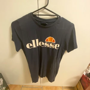 Mörkblå t-shirt från Ellesse - Snygg mörkblå t-shirt från Ellesse med deras ikoniska logga i vitt och orange på bröstet. Perfekt för en avslappnad stil. T-shirten har en klassisk passform och korta ärmar.