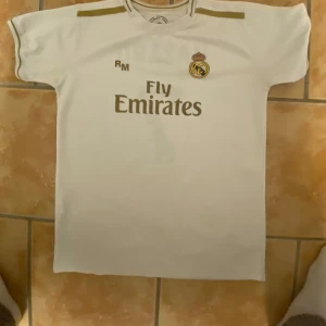 Vit Real Madrid fotbollströja - Säljer en vit Real Madrid fotbollströja från Adidas med korta ärmar. Tröjan har klubbens emblem och Fly Emirates-logga på framsidan. På baksidan finns namnet 'Hazard' och nummer 7 i guld. Perfekt för fans av laget!