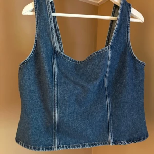 Jeanslinne med dragkedja - Säljer ett snyggt jeanslinne i blått med dragkedja bak. Linnet har en stilren design med breda axelband och rak u-ringning. Helt ny med prislapp kvar.